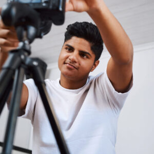 cropped-shot-of-a-young-man-using-a-camera-on-a-tr-2023-01-10-23-01-19-utc Cropped shot of a young man using a camera on a tripod to record himself at home.